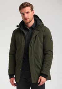 Giacca isolante verde oliva con cappuccio, zip frontale, bottoni a pressione e uno strato interno imbottito nero con un motivo geometrico.