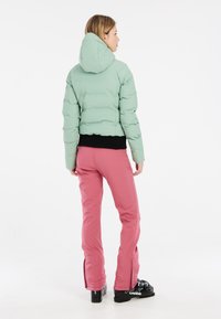 Lichtgroene donsjas met capuchon, gecombineerd met roze skibroeken met ritssluitingen aan de onderkant en zwarte skibotten. Soepele, gewatteerde textuur.