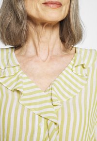 Femme mûre aux cheveux gris mi-longs portant un chemisier rayé jaune et blanc avec un col à volants, visible du cou à la poitrine.