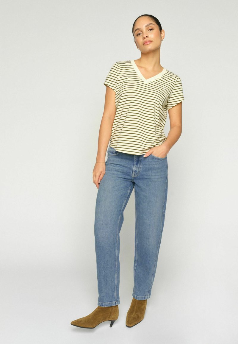 Woman standing wearing a short-sleeve striped V-neck top, blue jeans, and brown ankle boots against a plain light background.