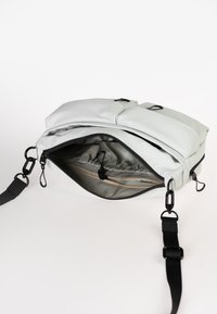 Helle graue Schultertasche mit weicher Textur, mit einem Reißverschlussöffnung, Innenfach und verstellbarem schwarzen Tragegurt. Zwei äußere Schnallen sind sichtbar.