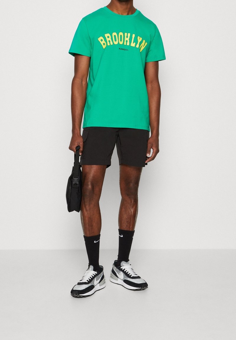 Hombre vistiendo una camiseta verde de "Brooklyn", pantalones cortos negros, calcetines negros de Nike, zapatillas en blanco y negro, sosteniendo una pequeña bolsa negra a su lado.