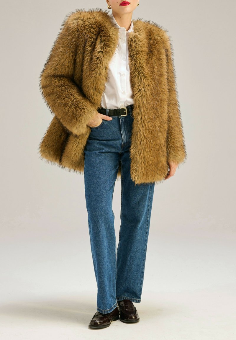 Futrzana kurtka w jasnobrązowym kolorze, w oversize'owym fasonie z wysokim kołnierzem, noszona na białą koszulę i niebieskie jeansy z wysokim stanem, w zestawieniu z ciemnymi butami.
