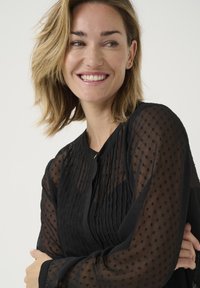 Schwarze Bluse aus transparenter Stoff mit strukturierten Punkten und Faltendetails auf der Vorderseite. Runder Ausschnitt und Knopfverschluss.