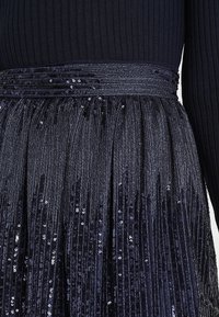 Jupe en sequins bleu marine avec un design texturé à niveaux. Dotée d'une taille brillante, accentuée par des sequins horizontaux et des franges verticales.