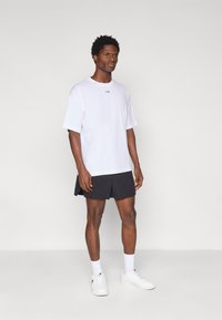 Λευκό κοντομάνικο μπλουζάκι με μικρό λογότυπο, συνδυασμένο με μαύρο σορτς και λευκά αθλητικά παπούτσια. Το ύφασμα φαίνεται λείο και ελαφρύ.