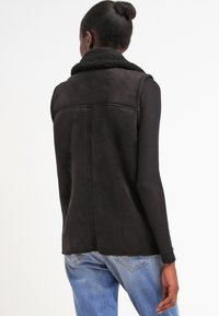 Donna che indossa un gilet in shearling nero senza maniche sopra una maglietta nera a maniche lunghe e jeans blu, capelli raccolti a chignon, in piedi di spalle.