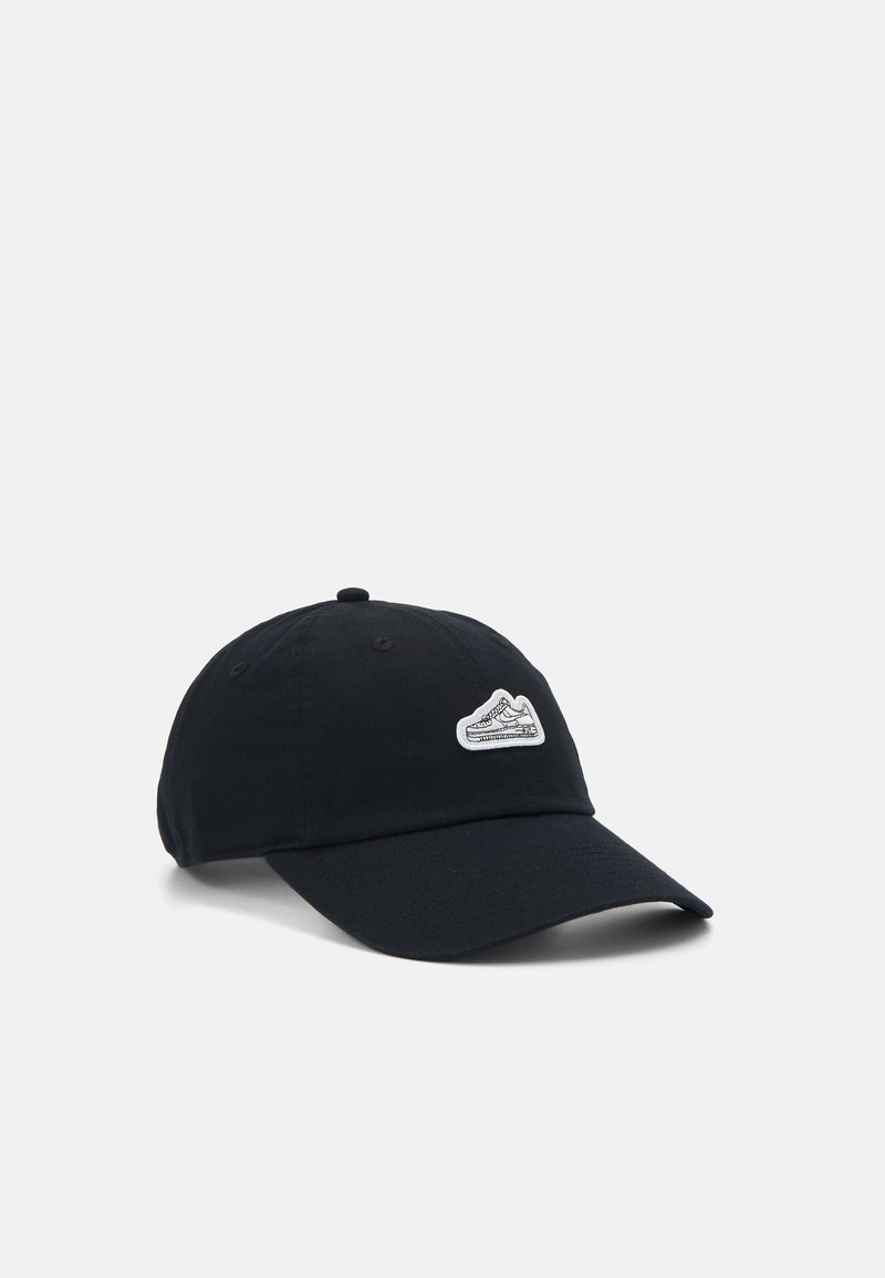 Μαύρο καπέλο από βαμβάκι με καμπύλη γείσο και λευκή ετικέτα με αθλητικά παπούτσια στο εμπρός μέρος. Ρυθμιζόμενο λουράκι στην πλάτη για ασφαλή εφαρμογή.