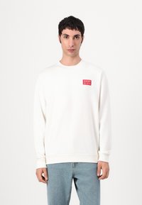 Hvit sweatshirt med ribbede mansjetter og nederkant, med en rød firkantet logo på venstre bryst. Bært med lys blå denimjeans.