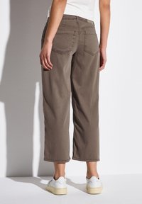 Pantalones de pierna ancha marrones de algodón, con dos bolsillos traseros y una cintura de altura media, combinados con zapatillas blancas sobre un fondo claro.