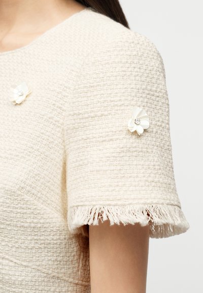 Κοντομάνικο κρεμ τοπ με υφή, διακοσμημένο με φράντζα και μικρά λευκά λουλούδια με κέντρα από κρύσταλλο στο μανίκι και το στήθος.