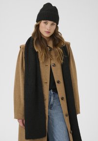 Brauner Wollmantel mit großen Knöpfen, kombiniert mit einem schwarzen Strickschal und einer Mütze. Der Mantel hat breite Schultern und eine entspannte Passform.
