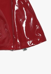 Czerwona, błyszcząca spódnica z winylu o gładkiej teksturze, z widocznym szwem i zamkiem błyskawicznym na dole. Dodatki obejmują metkę materiałową w pobliżu zamka.