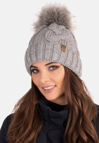 Berretto in maglia grigia con un motivo a treccia, decorato con un soffice pom-pom grigio sulla parte superiore. Presenta un piccolo patch in pelle marrone come accento.