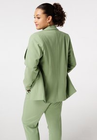 Groen op maat gemaakte pak met een gestructureerde blazer en bijpassende broek. De blazer heeft een achterpaneel; de stof heeft een gladde textuur.