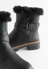 Schwarze Knöchelstiefel mit strukturierter Oberfläche, Kunstfellbund, Riemen mit Metallschnalle und kontrastierender brauner Sohle. Klobige, profiliierte Laufsohle.