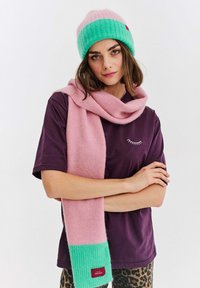 Pletená čepice se světle růžovou korunou a zeleným lemem; měkký růžový šál se zeleným akcentem; nošeno přes fialovou košili; uvolněný střih a pohodlná textura.