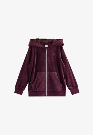Ragazza giovane in piedi e sorridente, indossa una felpa con cappuccio in velluto viola scuro e pantaloni abbinati con sneakers multicolori.
