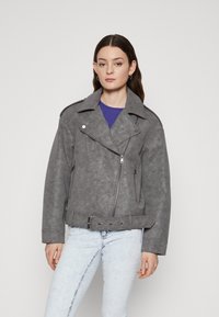 Veste de moto en faux suède gris avec des revers larges, une fermeture éclair asymétrique, des poches latérales et une taille ceinturée. Un jean bleu clair complète le look.