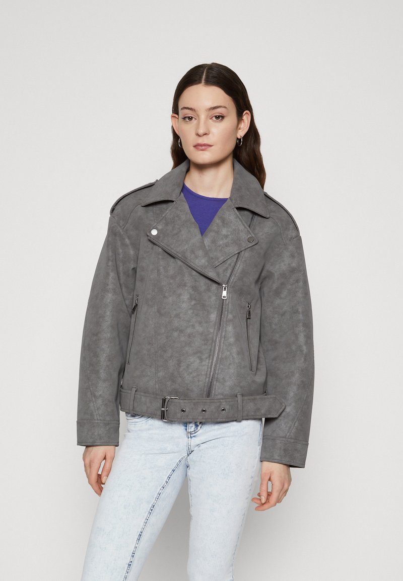 Veste de moto en faux suède gris avec des revers larges, une fermeture éclair asymétrique, des poches latérales et une taille ceinturée. Un jean bleu clair complète le look.