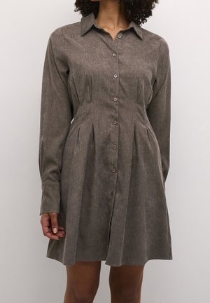 Donna che indossa un vestito camicia marrone a costine con maniche lunghe e bottoni, con pieghe in vita, su uno sfondo semplice.