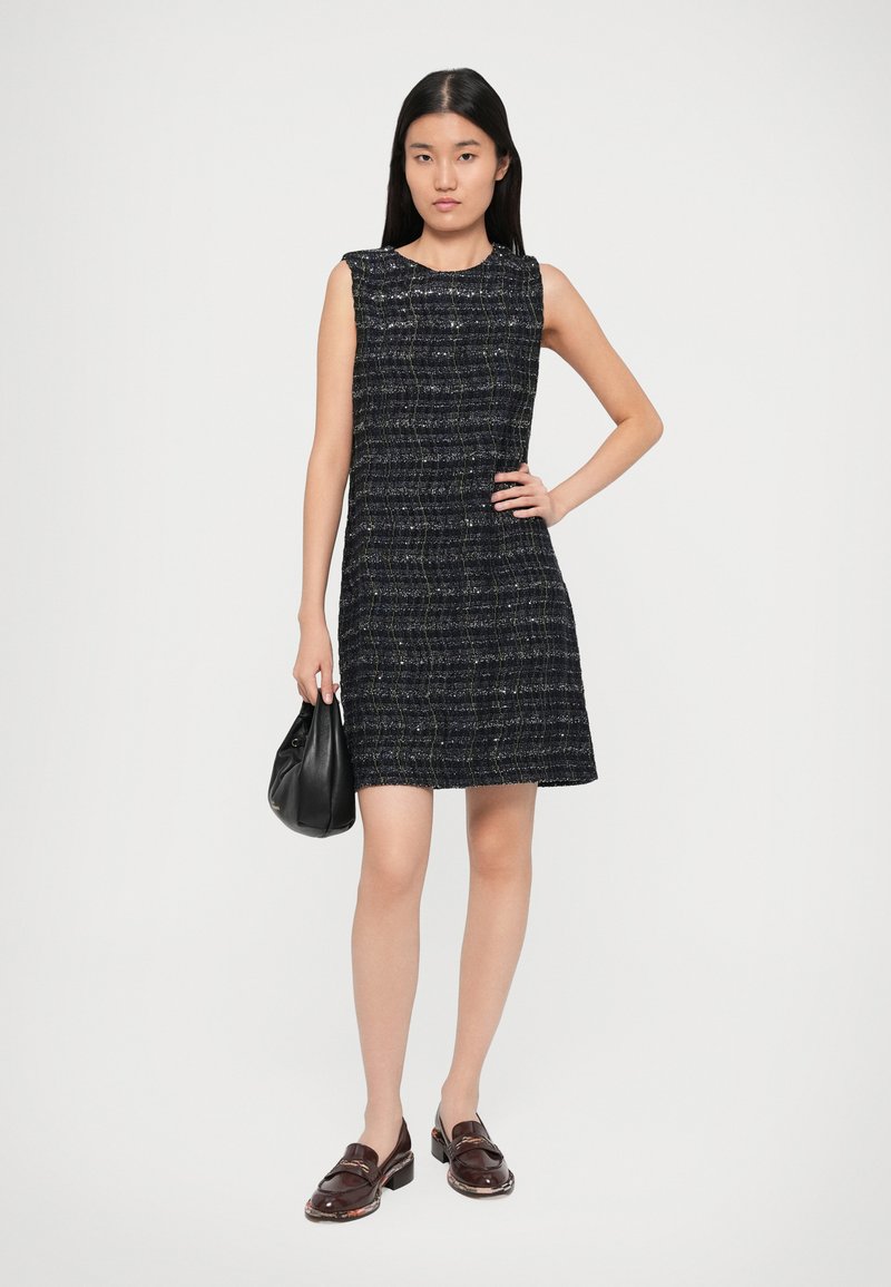 Marineblaues ärmelloses Kleid aus strukturiertem Stoff mit einem Karomuster. Das Modell hält eine schwarze Tasche und trägt dunkle Slipper.