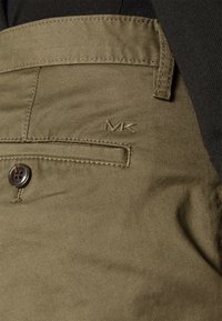 Olivově zelené kalhoty s texturovaným povrchem, s dekorativním knoflíkem a vyšitým logem "MK" blízko kapsy.