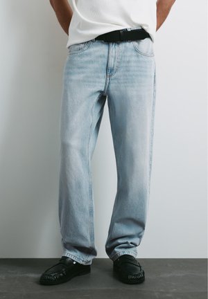 Personne portant un jean bleu clair à jambes droites, une ceinture noire, une chemise blanche et des chaussures noires, debout contre un mur clair uni.