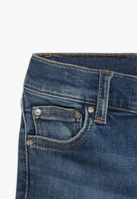 Des jeans en denim bleu foncé présentent un design classique à cinq poches, avec des coutures visibles, une petite poche avant et des rivets métalliques.