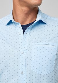 Camicia button-up azzurro chiaro con un motivo a design di piccoli quadrati. Presenta una tasca e un tessuto morbido e testurizzato.