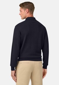 Jūras spēku zils adīts džemperis ar garām piedurknēm un apkakli, ar ribotu piedurkņu un apakšmalu apdari, saskaņots ar gaiši bēšām biksēm.