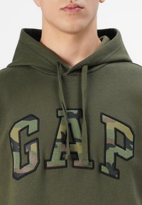 Egy személy közeli képe, aki olajzöld kapucnis felsőt visel, a mellkasán nagy, camouflázs mintázatú "GAP" felirattal, és a zsinórok lógva.