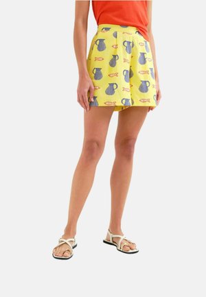 Mujer con pantalones cortos amarillos con estampados de jarra azul y peces naranja, una camiseta naranja y sandalias blancas con tiras, de pie contra un fondo blanco.