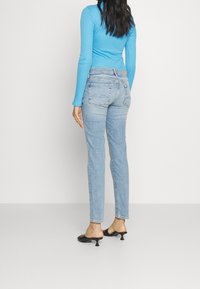 Los jeans de tiro alto azul claro presentan un corte recto, dos bolsillos traseros y un sutil desgaste. Se combinan con una camiseta ajustada de manga larga azul y zapatos negros.