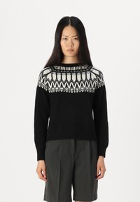 Schwarzer Strickpullover mit einem gemusterten weißen und schwarzen Schulterbereich. Lange Ärmel, gerippter Ausschnitt und entspannter Schnitt. Kombiniert mit grauen Shorts.
