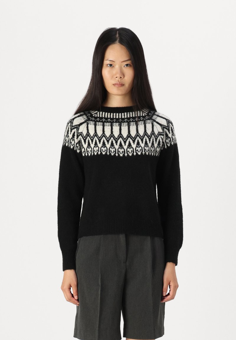 Schwarzer Strickpullover mit einem gemusterten weißen und schwarzen Schulterbereich. Lange Ärmel, gerippter Ausschnitt und entspannter Schnitt. Kombiniert mit grauen Shorts.