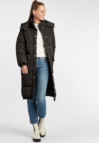 Svart puffjacka med hög krage, tryckknappar och elastiska ärmslut. Bärs över en krämfärgad tröja och ljusblå jeans med beige ankelstövlar.