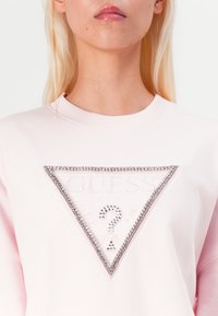 Pink sweatshirt featuring a triangular logo with embossed text and rhinestone accents. Soft fabric with a crew neckline and long sleeves.