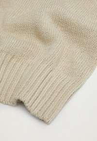 Beige gestrickter Stoff mit geripptem Bündchen, der eine weiche Textur und gefaltetes Aussehen zeigt. Dezente Farbvariationen sind sichtbar.