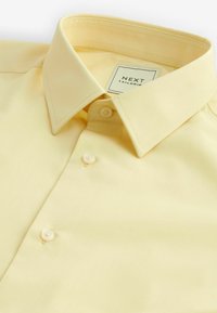 Chemise jaune clair à boutons avec un col pointu, deux boutons visibles et une étiquette indiquant «NEXT TAILORING» à l’intérieur du col.