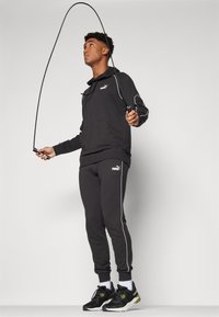 Black hooded sweatshirt with kangaroo pocket and white piping, paired with matching pants. The model is skipping with a black jump rope.