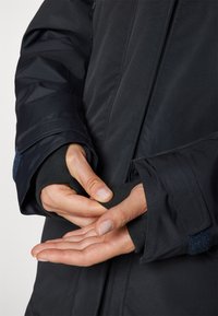Zwarte geïsoleerde jas met een gestructureerde afwerking, voorzien van duimopeningen in de mouwen, verstelbare manchetten en contrasterende binnenstof.