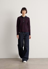 Woman with short black hair wears dark wide-leg jeans, white sneakers, and a black and red striped long-sleeve polo shirt, standing against white wall.
