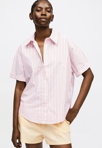 Kvinde iført en løs lyserød og hvid vertikalt stribet kortærmet skjorte med beige shorts, stående med den ene hånd i lommen mod en hvid baggrund.