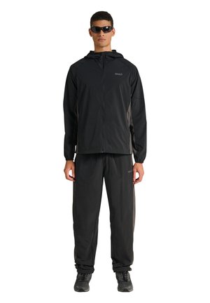 Mężczyzna ubrany w czarną sportową kurtkę z kapturem i dopasowane spodnie, czarne trampki oraz ciemne okulary przeciwsłoneczne, stojący na tle białego tła.