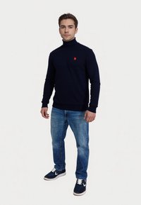 Joven vestido con un suéter de cuello alto azul marino con un logo rojo, jeans azules y zapatillas deportivas azules, de pie frente a un fondo blanco liso.