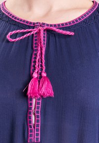 Marinblus med rund halsringning som har rosa geometrisk brodering, en framknäppning och flätade rosa tofsar vid knytningen.