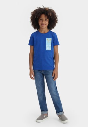 Enfant aux cheveux bouclés portant un t-shirt bleu "Suis tes rêves", un jean bleu retroussé et des chaussures grises sans lacets, debout devant un fond uni.
