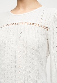 Λευκό μπλουζάκι από δαντέλα με σχέδιο eyelet και ανθισμένη διακοσμητική λεπτομέρεια στο στήθος, με φουσκωτά μακριά μανίκια, εμφανιζόμενο από τον λαιμό μέχρι τη μέση του κορμού.