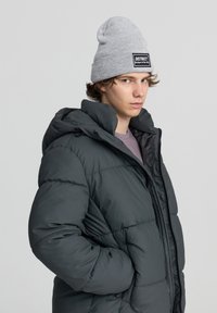 Czarna szara kurtka puchowa z wysokim kołnierzem i zamkiem błyskawicznym z przodu, z przeszywanymi panelami i odpinanym kapturem. W zestawie z jasnoszarym czapką beanie.